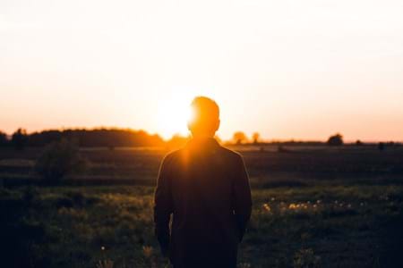 A man standing in an open field with the sun setting in front of him.