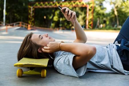 A teenager laying down on the ground resting his head on a yellow skateboard as he holds a phone in front of his face.
